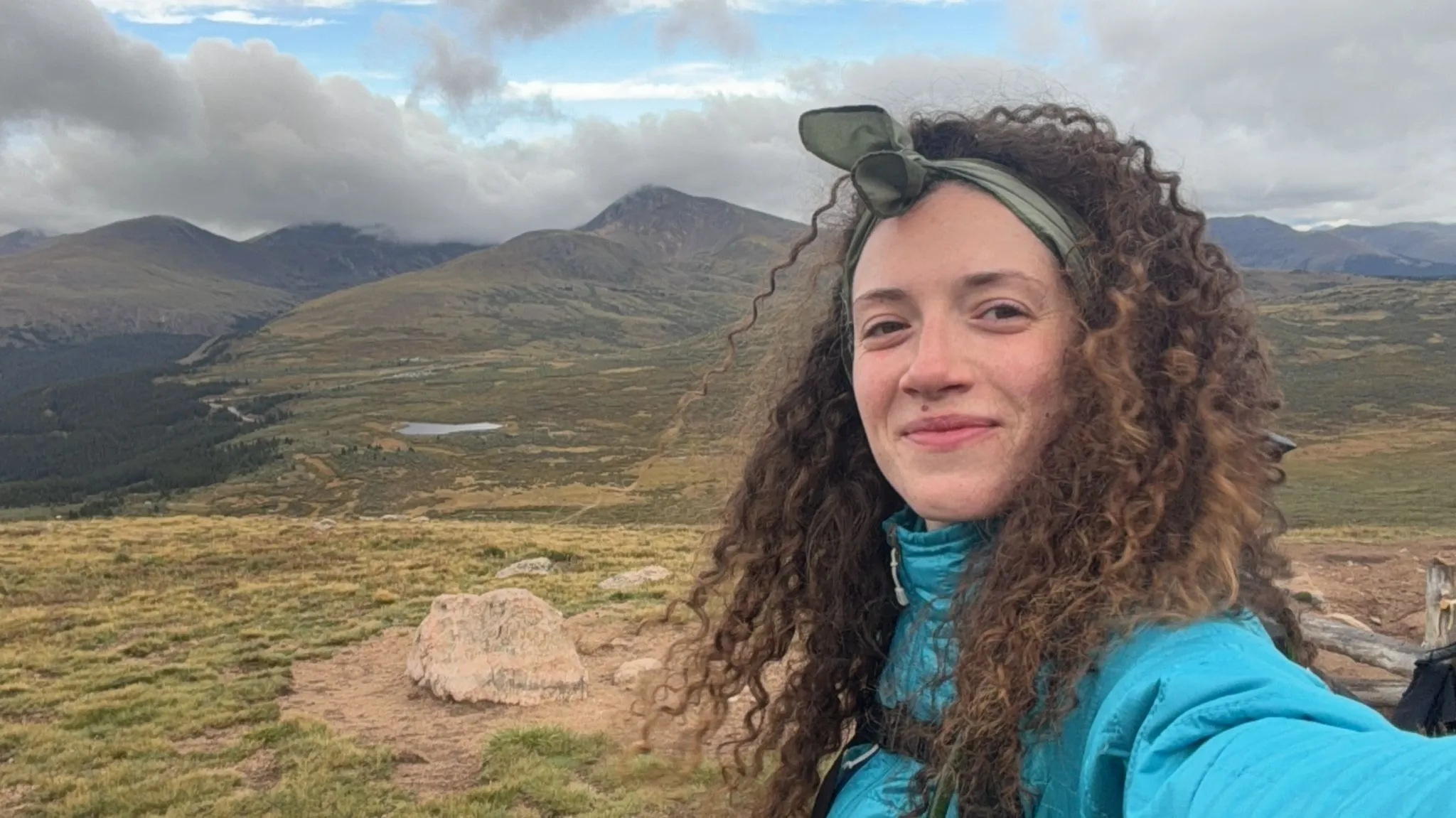 Dóra on a high ridge in a blue jacket, a lake and mountains behind her
