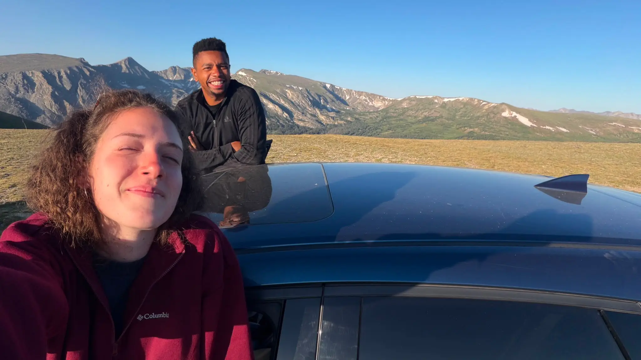 Dóra in the car and Leon standing behind it, mountains in the background